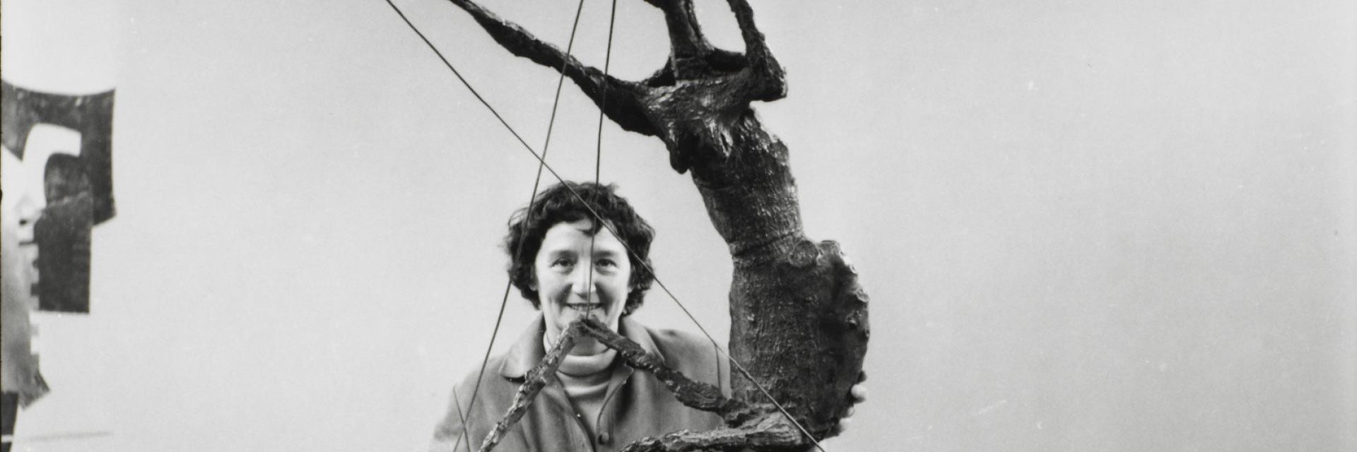 Photo e nnoir & blanc de Germaine Richier dans une salle d'exposition, face caméra et souriante, derrière une de ses sculptures