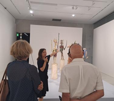 Dans une salle de l'exposition "Germaine Richier", une guide-conférencière tient un petit micro et montre une œuvre dans le fond à un groupe de visiteurs.