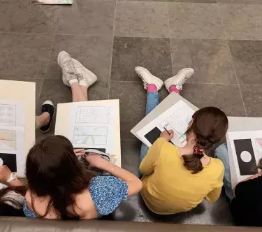 Vues de dessus, des jeunes filles sont assises, appuyées sur un mur, et dessinent sur des cahiers.