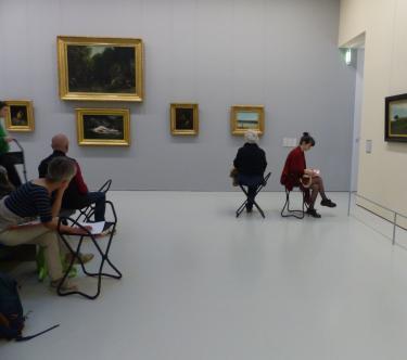 Dans une salle du musée, un groupe de gens assis sur des tabourets sont en train de prendre des notes.