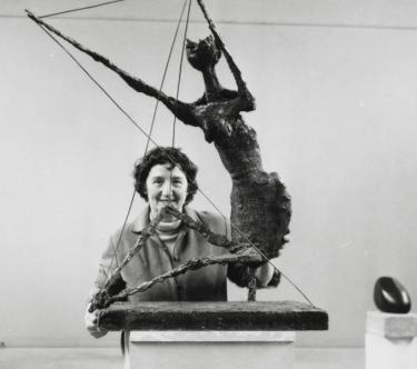 Photo en noir & blanc sur laquelle on peut voir Germaine Richier, souriante, derrière sa sculpture "La Fourmi" dans une salle d'exposition.