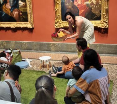 Devant le tableau "Vertumne et Pomone" de Jean Ranc, une médiatrice montre un panier de fruits factices à un groupe de parents et de bébés assis sur un tapis vert.