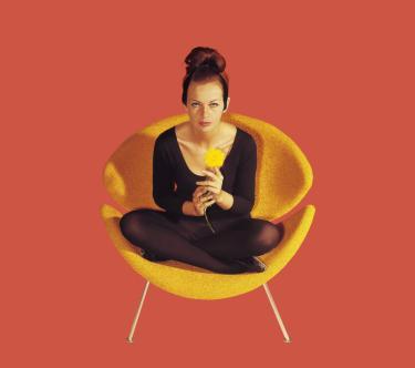 Une femme est assise en tailleur sur un fauteuil orange ; le tout détouré sur un fond uni corail.Une femme est assise en tailleur sur un fauteuil orange ; le tout sur un fond uni couleur corail.