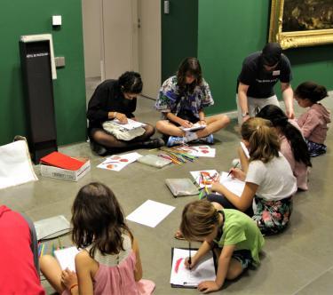 Un médiateur et un groupe d'enfants sont assis au sol dans une salle du musée. Les enfants dessinent avec des crayons de couleurs mis à disposition