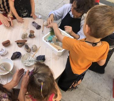 Cinq personnes (une adulte et quatre enfants) sont installés autour d'une table basse ronde ; ils pratiquent des arts manuels avec des pinceaux et divers petits objets