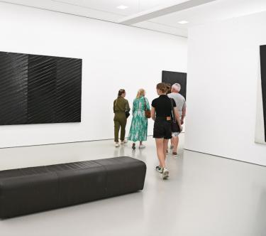 À gauche, un tableau de Pierre Soulages. À droite, un groupe de visiteurs passe derrière un mur. Au premier plan, un banc.