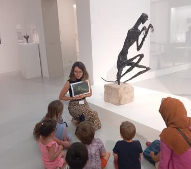 Dans une salle de l'exposition Germaine Richier, une médiatrice, de face, montre une image de mante religieuse à un groupe d'enfants et à une adulte, à côté d'une sculpture en bronze représentant une mante religieuse stylisée.