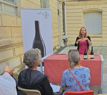 Devant une table, une vigneronne présente ses vins à du public.