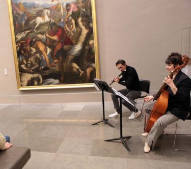 Dans une salle de musée, deux musiciens jouent du viloncelle et de la flûte devant le public.