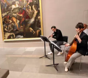 Dans une salle du musée, on voit, sur la droite, deux musiciens assis : un homme qui joue de la flûte traversière et une femme qui joue de la viole de gambe. Sur la gauche, une femme plutôt âgée les regarde.