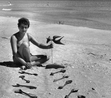 Une jeune femme (Valentine Schlegel) est assise sur la plage et présente plusieurs sculptures en forme d'ustensiles de cuisine ; en fond, deux baigneurs dans la mer.