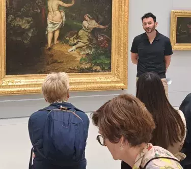 Un médiateur fait face à un groupe de vsiiteurs, assis, devant le tableau "Les Baigneuses" de Gustave Courbet.